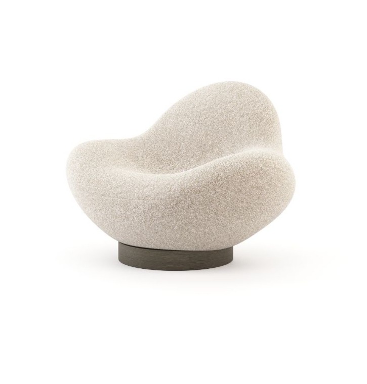 The Stone armchair, characterised by a soft, rounded silhouette and a textured beige upholstery, sits on a dark circular base in a minimalist setting, highlighting its sculptural form inspired by stone.