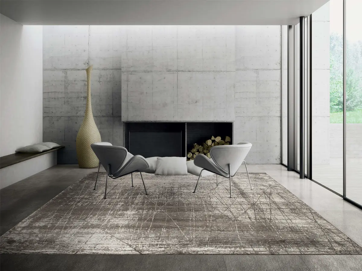 Foto of a rug with grey tones places in a living room with concrete walls, two chairs and a fireplace.