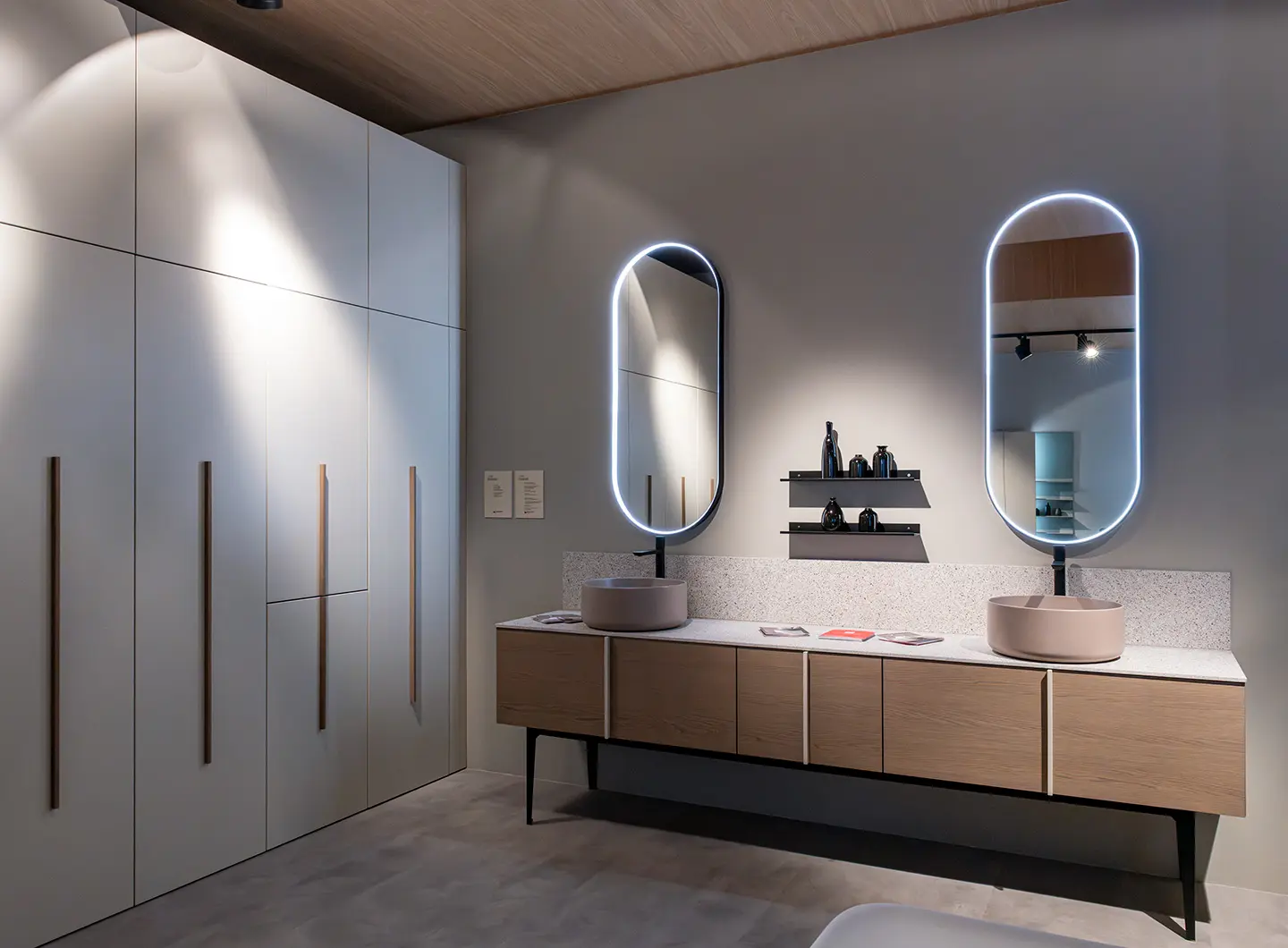 Light wood bathroom cabinet with two round ceramic countertop washbasins and two oval mirrors, with full-height white cabinetry on the side 