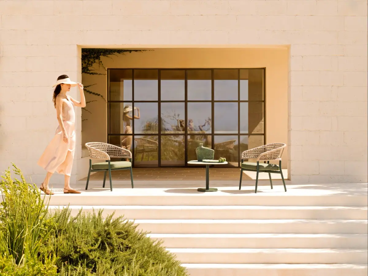 Patio esterno assolato con gradini bianchi, arredato con un tavolino basso e due poltroncine intrecciate di colore verde bosco della collezione Plinto di Varaschin. Sulla sinistra, una donna in abito chiaro e visiera parasole passeggia in relax.