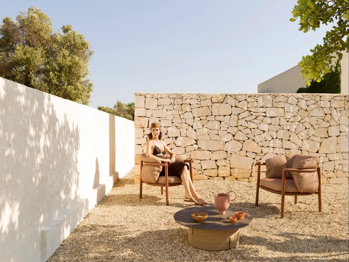 Due poltrone da esterno della collezione Allegra di Varaschin color terracotta, ambientati in un cortile assolato con ghiaia e muretti a secco bianchi. Una donna è seduta in relax su una delle poltrone.