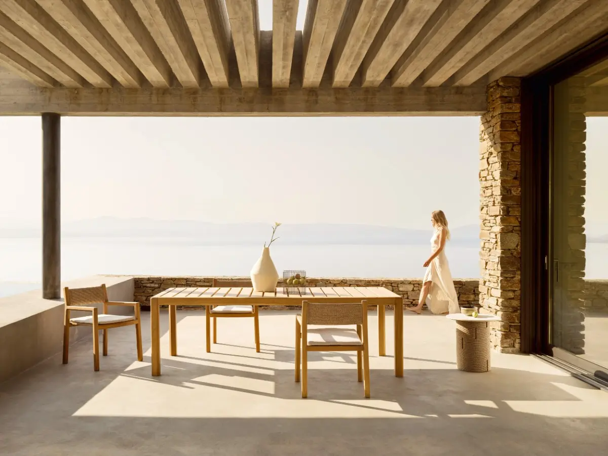 Grande terrazza panoramica all'aperto con pergola in cemento a vista sul mare. Al centro, il tavolo da pranzo rettangolare in legno di iroko della collezione Gianna di Varaschin con tre sedie abbinate. Sullo sfondo a destra, una donna in abito bianco chiaro passeggia vicino a un muro in pietra.
