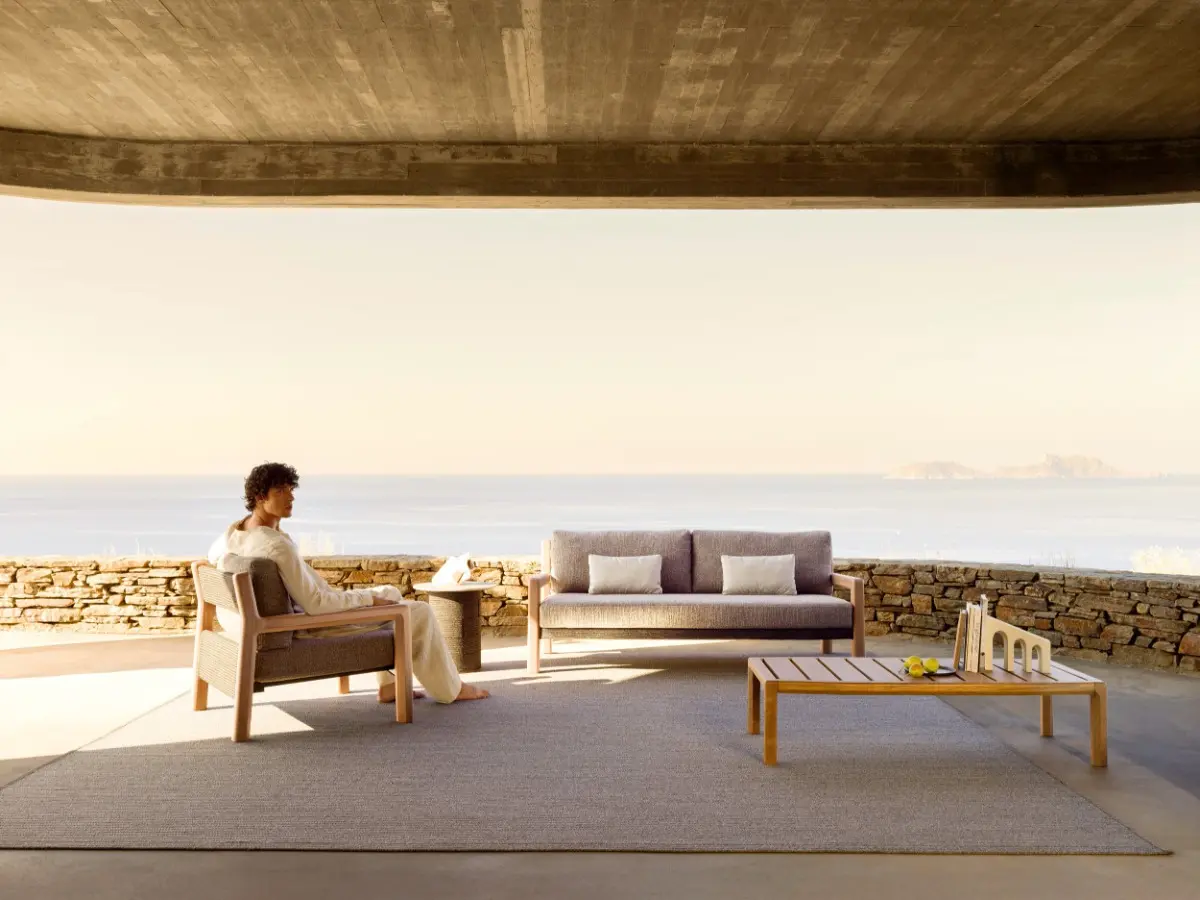 Area lounge all'aperto con vista mare, arredata con poltrona, divano e tavolino basso in legno di iroko della collezione Gianna di Varaschin. Una persona è seduta in relax sulla poltrona, posizionata su un grande tappeto sotto un porticato in cemento delineato da un muretto in pietra.