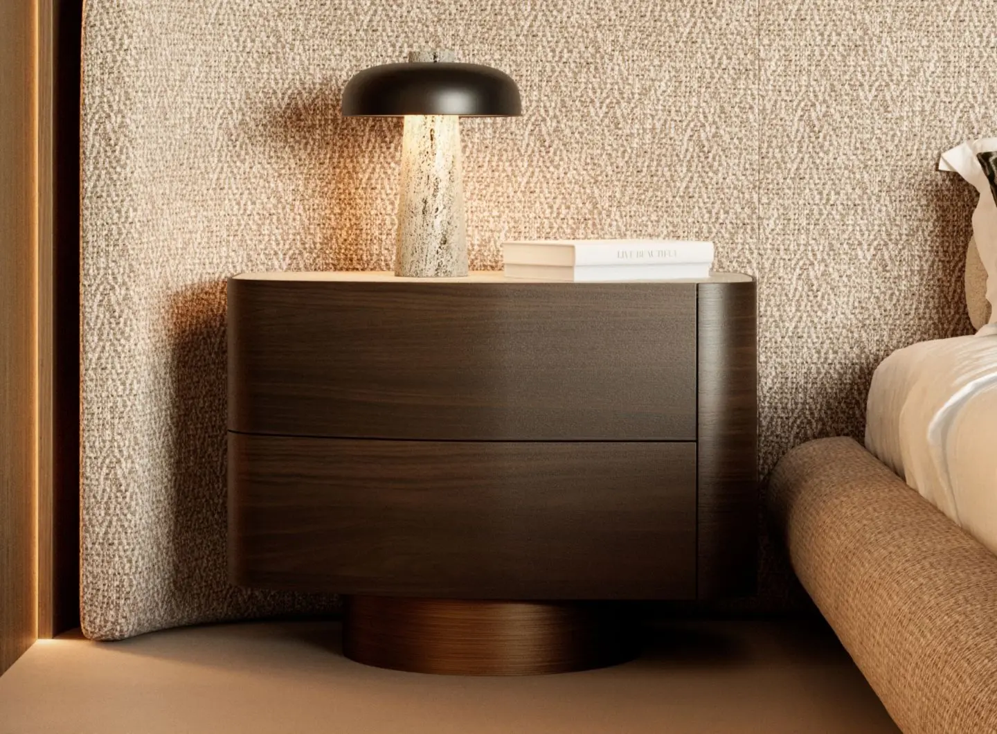 The Stone Night Table II in dark wood with a rounded silhouette and two drawers is styled with a stone‑textured lamp and stacked books, placed beside a beige upholstered bed against a subtly patterned wall.