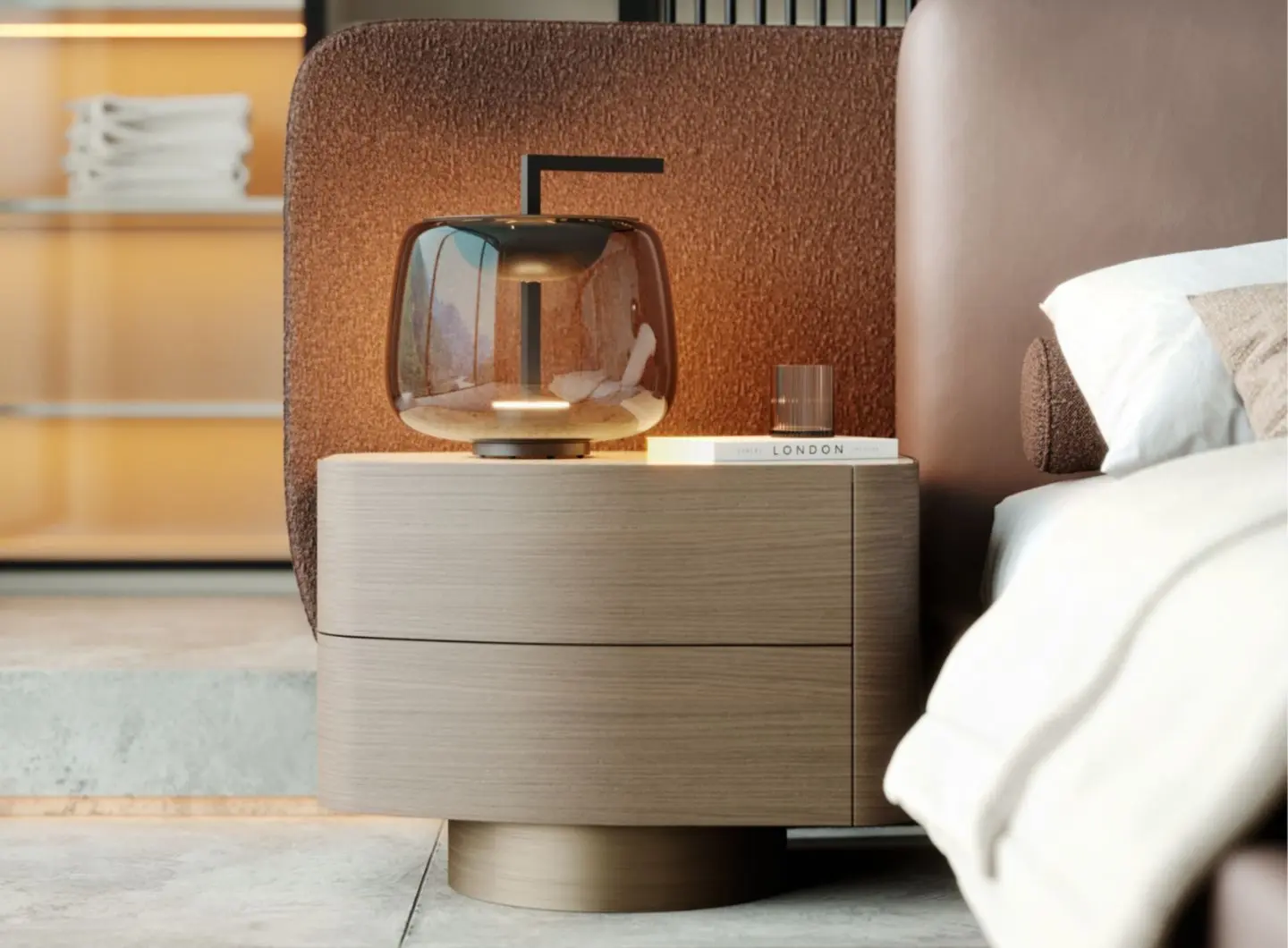 The Stone Night Table II in light wood with rounded edges and two drawers sits beside a bed with a brown upholstered headboard, styled with a glass lamp, a ribbed tumbler, and a book on its surface in a warm, modern bedroom.