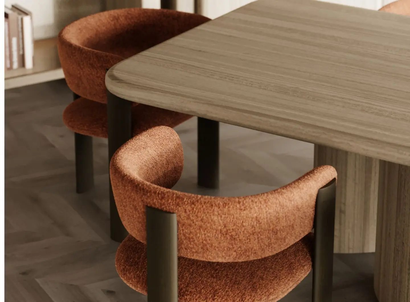 The Dolomites Chair is shown in a dining setting as a rust‑colored upholstered chair with a curved backrest and cushioned seat, supported by dark cylindrical legs, placed beside a light wood table with books on the shelf behind it.