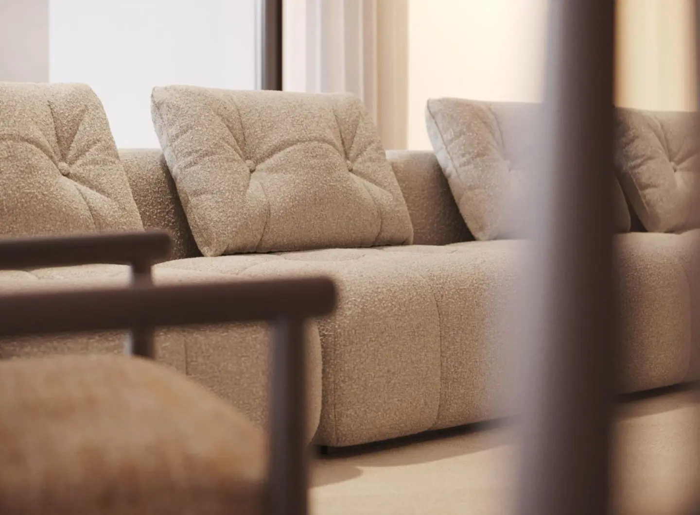 The Antónia Sofa is shown in a close-up view as a beige bouclé modular seat with deep tufted cushions and soft, textured upholstery, accompanied by a matching armchair with wooden armrests in the foreground, all set within a warm, neutral-toned interior that highlights the sofa’s tactile richness and minimalist design.