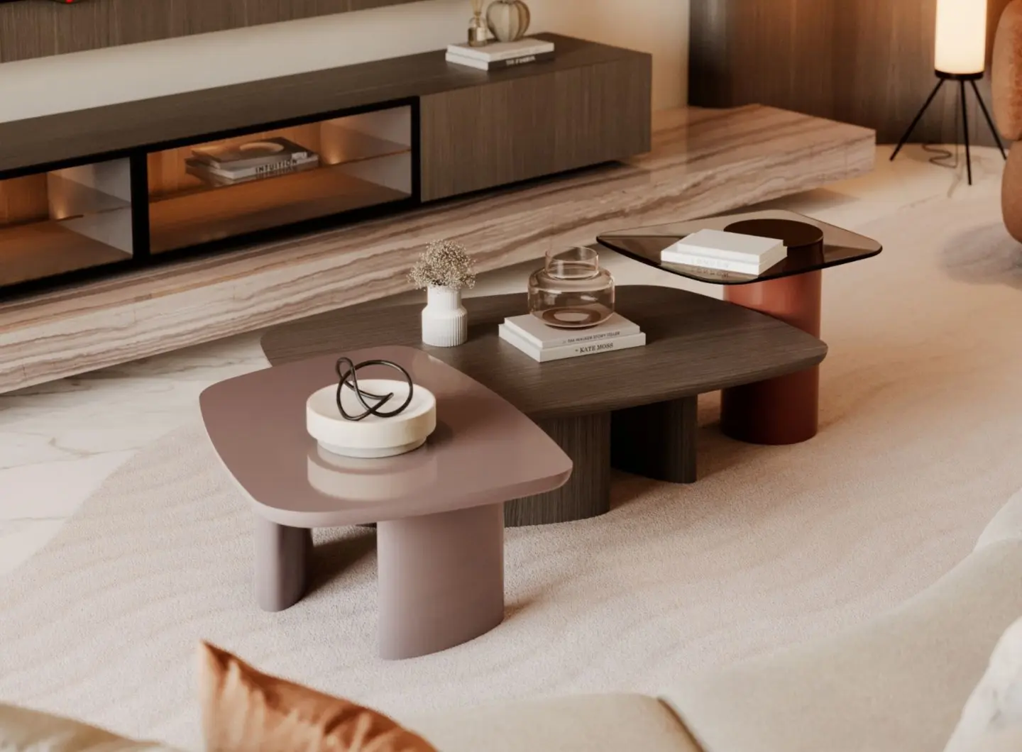 The Dolomites Coffee Table is presented as a set of three rounded tables with mixed materials, including a mauve‑toned top, a dark wood top, and a tinted glass top supported by a copper‑colored cylindrical base, arranged on a light rug with decorative objects and books nearby, featuring brass accents within the table bases.