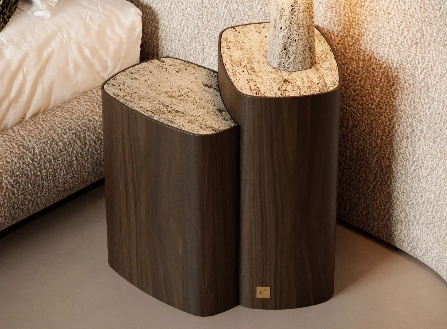 The Dolomites Side Table is shown as a pair of wooden tables with an organic curved base and polished stone tops in beige and brown tones, placed beside a bed, with one table holding a cylindrical stone object and a small brass plaque visible near the base.