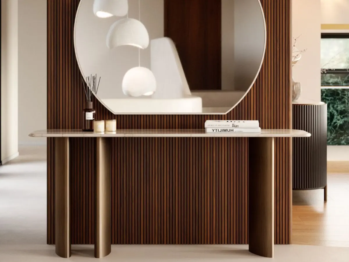 The Dolomites Console appears against a wall with vertical wooden slats, featuring a rounded stone‑like top and dark cylindrical legs, styled with candles, books, and a diffuser beneath a large circular mirror that reflects the surrounding interior.