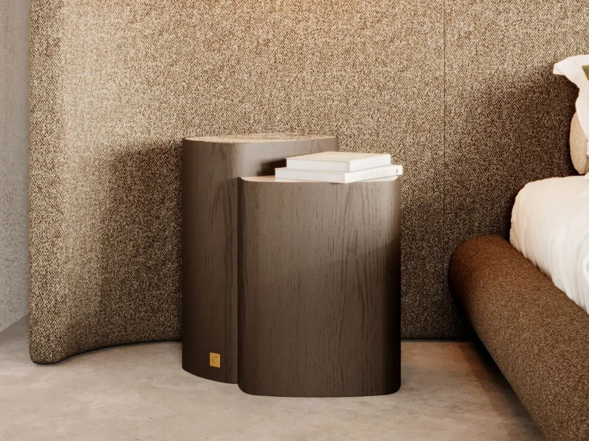 The Dolomites Side Table appears as two cylindrical wooden tables with an organic, softly rounded form in a dark finish, placed beside a bed, with the taller table holding two stacked books against a textured beige wall panel in a warm minimalist bedroom.
