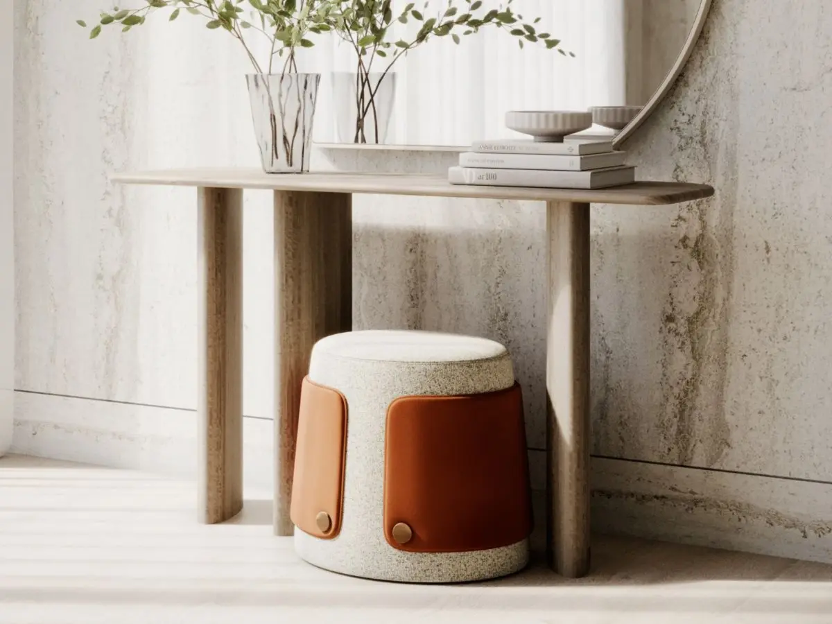 The Dolomites Stool appears beneath a wooden console table as a cylindrical seat with an organic rounded form, upholstered in textured beige fabric with brown leather side panels fastened by round metallic details, placed in a minimalist interior with natural materials.