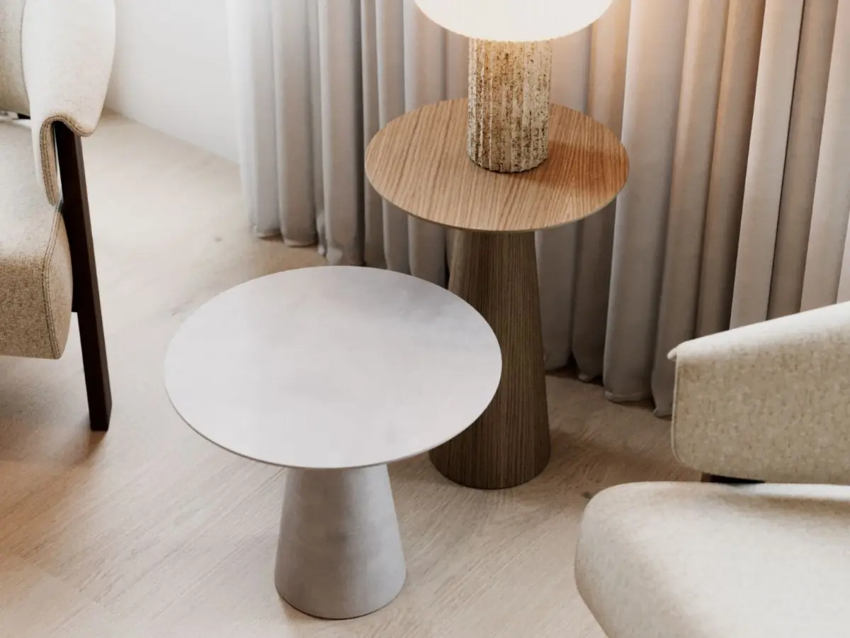 The Olivier Side Tables appear side by side in a minimalist interior, with the grey table in the foreground and the wooden table supporting a textured lamp, set against soft curtains and neutral furnishings that emphasize their sculptural curves and quiet Nordic‑inspired elegance.