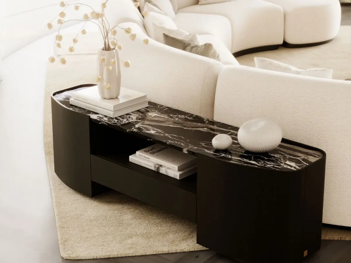 The Stone low console table, with a polished black stone top and a rounded dark wood base, is positioned behind a cream-coloured curved sofa, adorned with a white vase, sculptural decorative pieces and stacked books, in a living room decorated in soft, neutral tones.