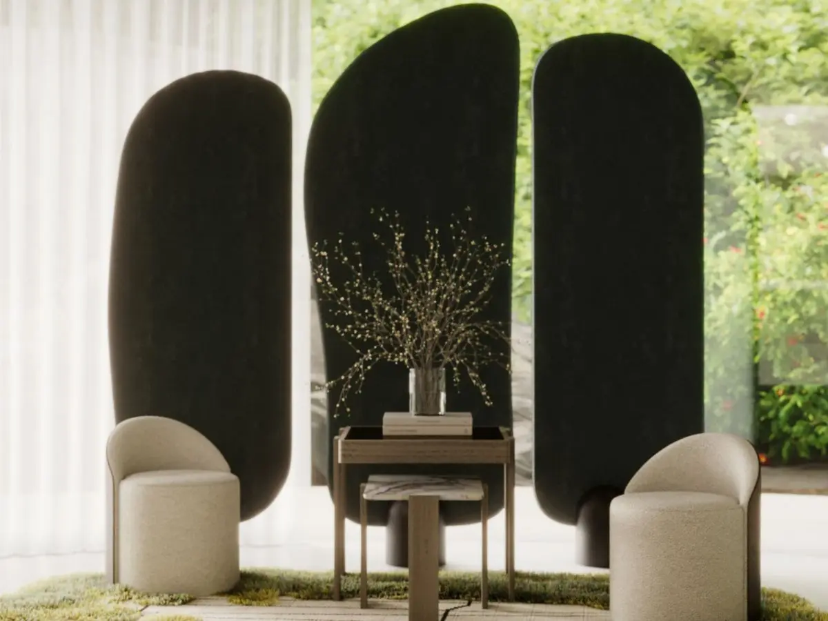 The Stone Screen appears as three large dark sculptural panels with smooth, organic curves, positioned behind two rounded beige chairs and a wooden table in a softly lit room with sheer curtains and greenery visible outdoors.