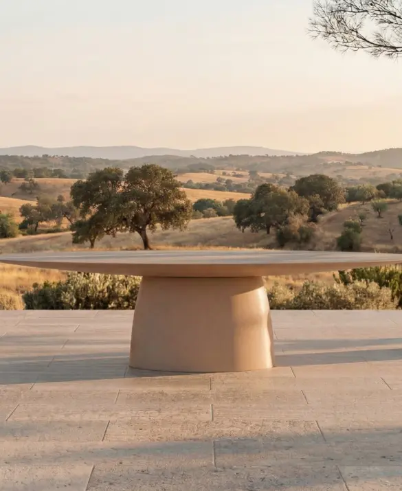 MAGNA Dining Table by GANSK on an outdoor terrace, a large round table with a sculptural pedestal base and two chairs overlooking a countryside landscape.