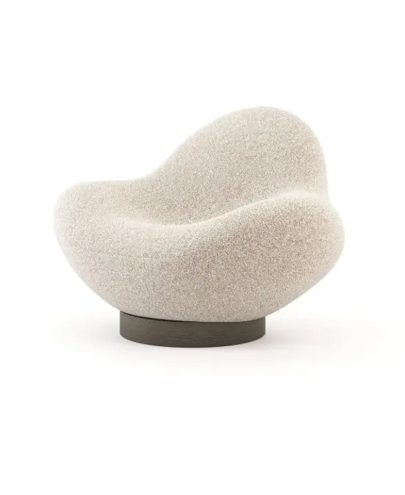 The Stone armchair, characterised by a soft, rounded silhouette and a textured beige upholstery, sits on a dark circular base in a minimalist setting, highlighting its sculptural form inspired by stone.
