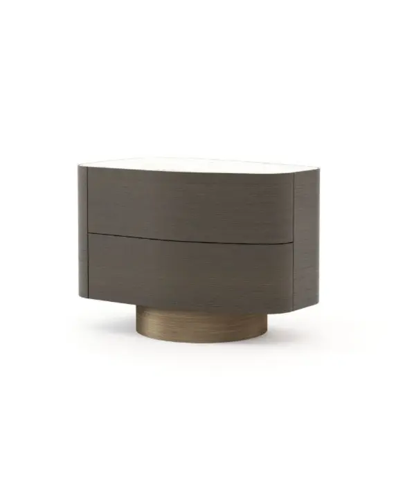The Stone Night Table, crafted from dark wood with two rounded drawers and a smooth white stone top, rests on a circular base in brushed bronze and is displayed in a minimalist setting that highlights its oval silhouette.