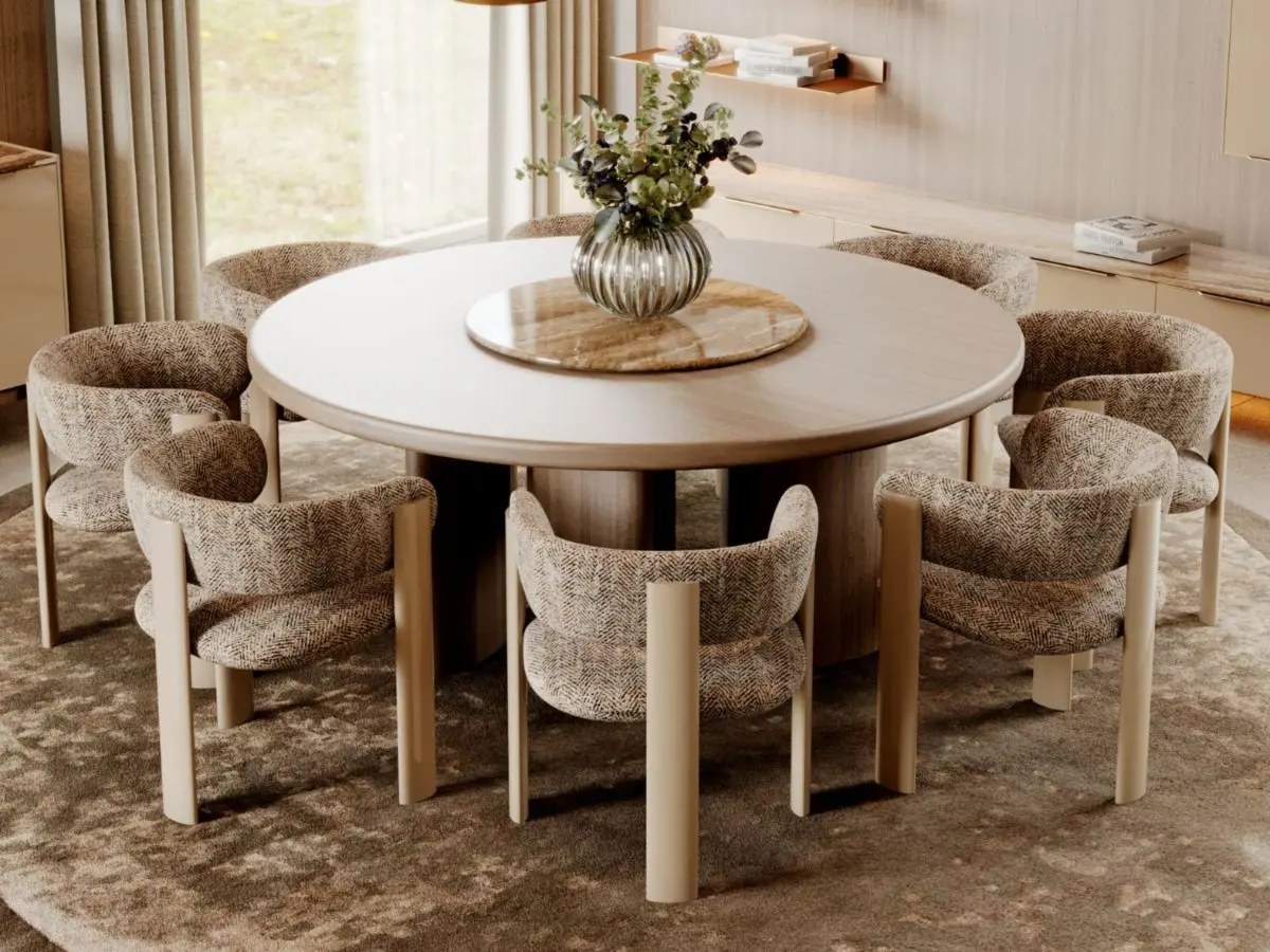 The Dolomites Round Dining Table is shown in an elegant dining area with six textured upholstered chairs arranged around it, set on a soft patterned rug. A floral centerpiece sits atop the table, complemented by beige curtains, built‑in wooden shelving, and a warm, contemporary ambiance.