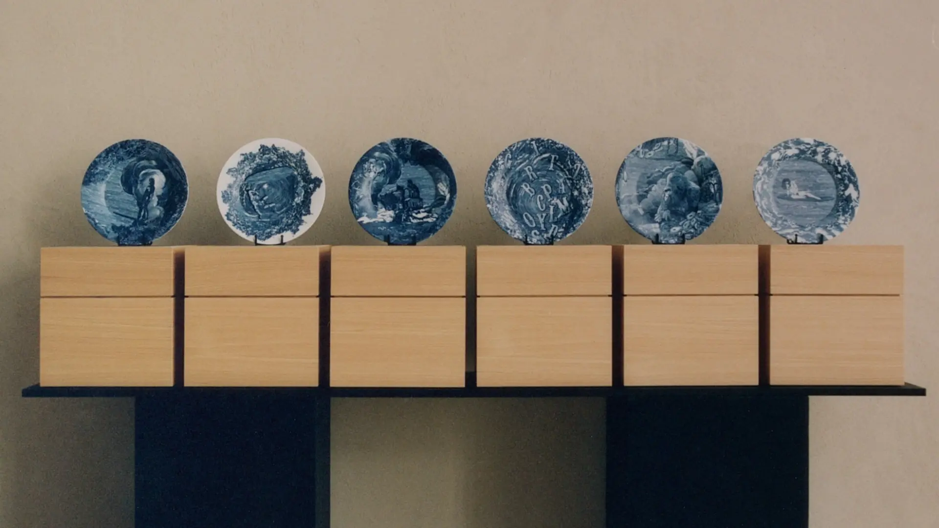 Serie di sei piatti della collezione Equinox I, disposti in fila sopra cassette di legno minimaliste.