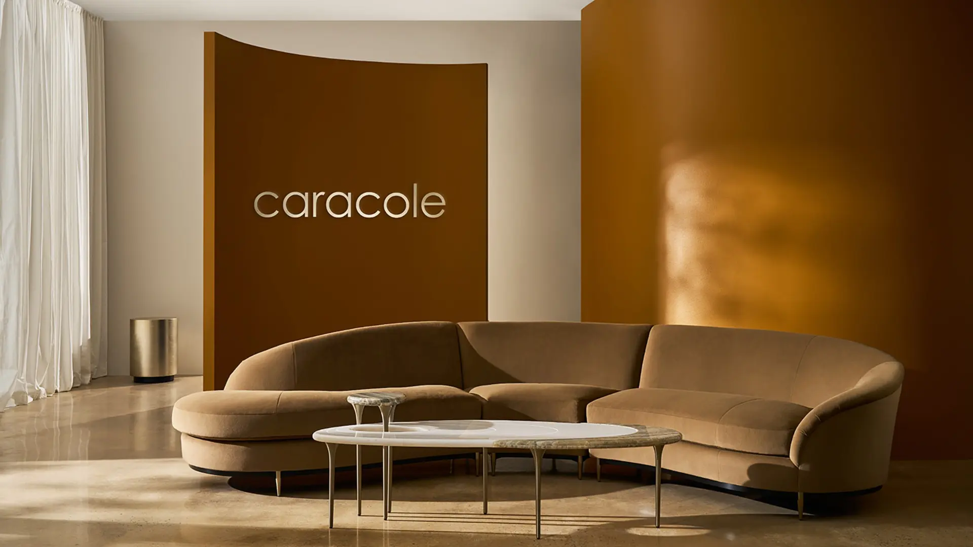 Caracole luxury furniture showroom featuring a curved, camel-colored sectional sofa with elegant marble coffee tables. The modern design blends warm ochre tones with minimalist decor, showcasing Caracole's commitment to sophisticated, timeless interiors. Sunlight highlights the polished concrete flooring, enhancing the brand's signature style of casual elegance.